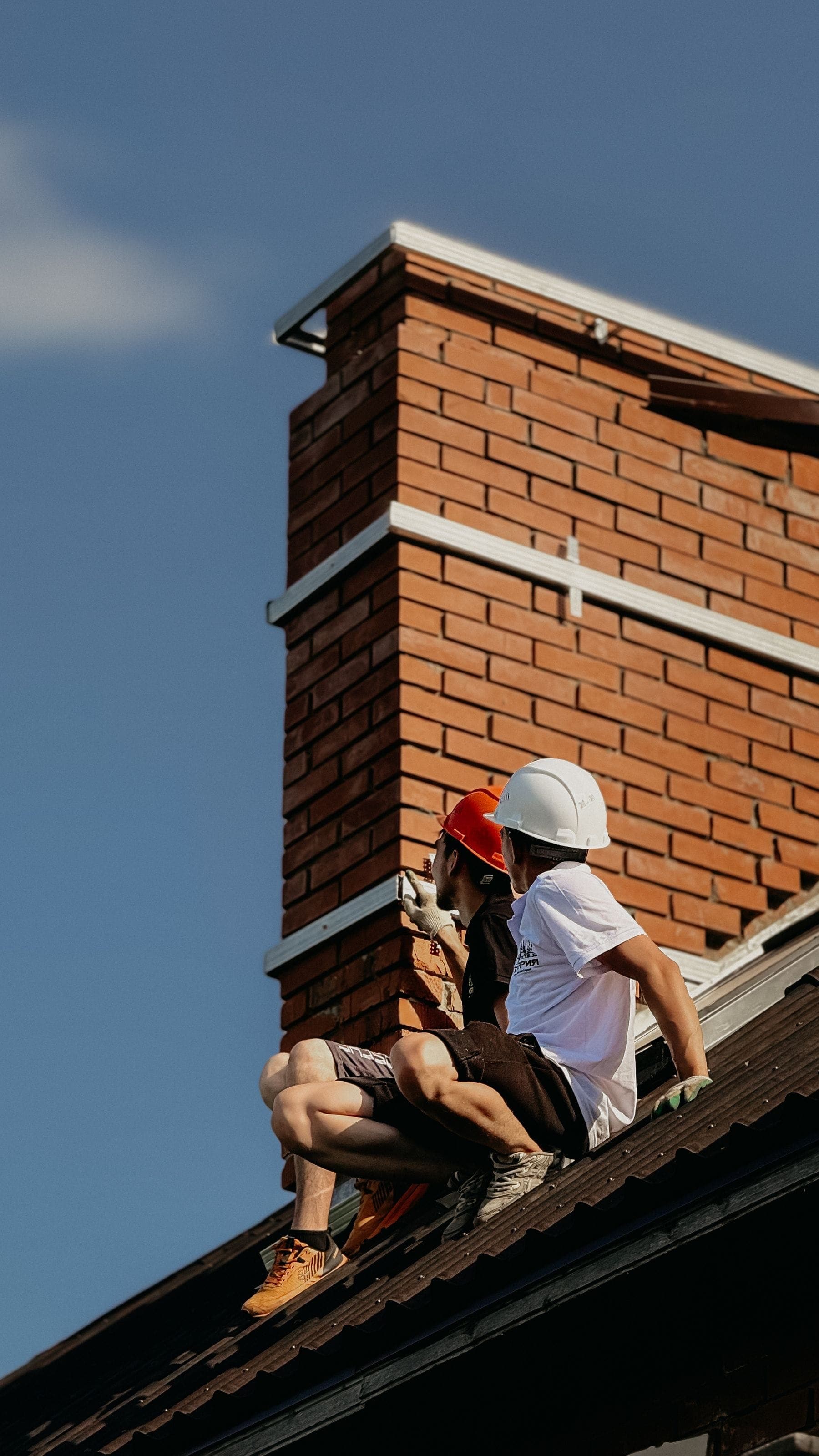 Two roofers working at the base of a red-brick chimney stack